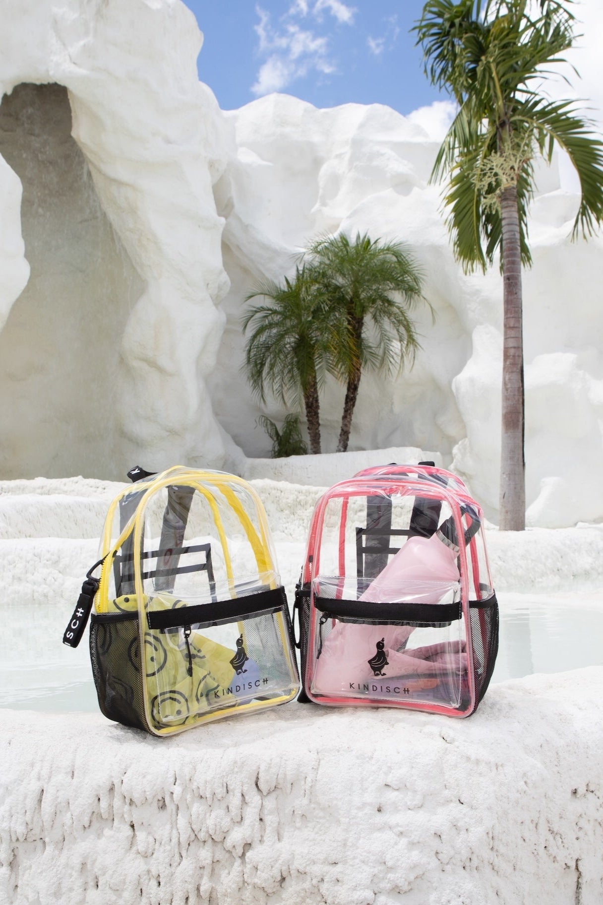 A KINDISCH Kids Backpack - Pink from kindischworld.com rests by a white stone pool, surrounded by palm trees and rocks, its spacious exterior pocket ready to hold swim gear under the bright blue sky.