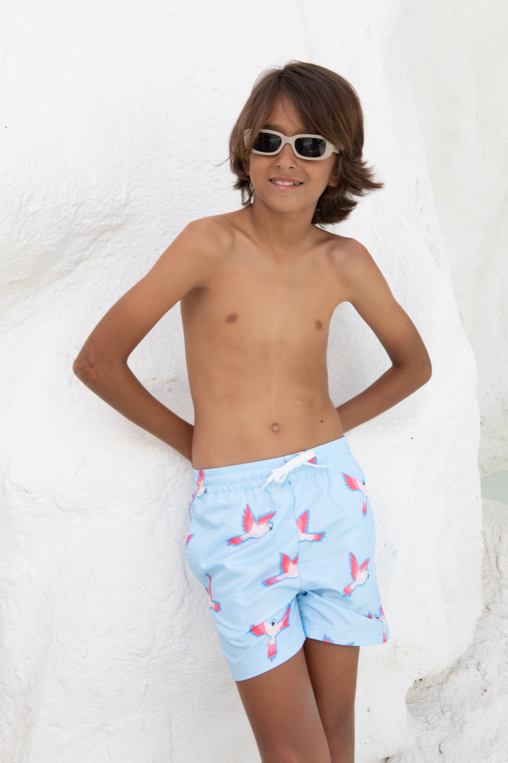 Ein Junge mit braunen Haaren trägt die Badehose PARROT von kindischworld.com aus recyceltem Polyester mit pinkem Papagei-Print, steht lässig vor einer weißen Wand und lächelt mit Sonnenbrille, die Hände hinter dem Rücken.