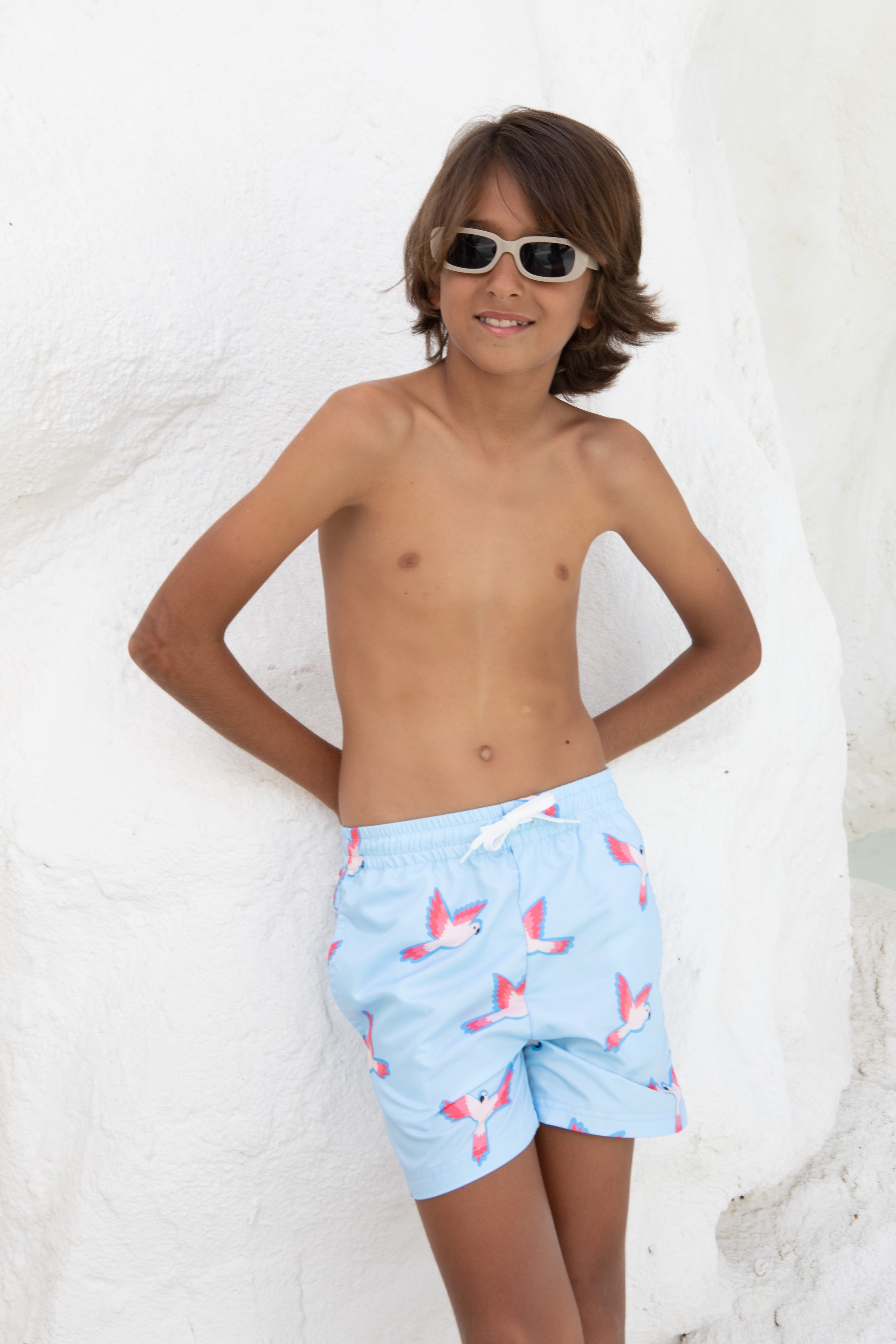 Ein Junge mit braunen Haaren trägt die Badehose PARROT von kindischworld.com aus recyceltem Polyester mit pinkem Papagei-Print, steht lässig vor einer weißen Wand und lächelt mit Sonnenbrille, die Hände hinter dem Rücken.
