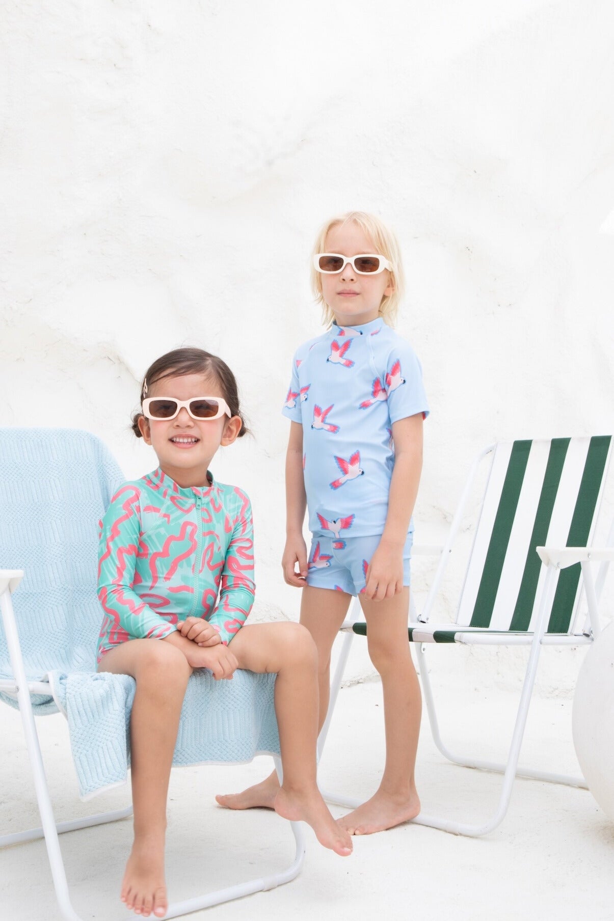 A boy wears the colorful Boys 2-piece - PARROT swimwear from kindischworld.com, made of recycled polyester. He stands beside a girl, both barefoot by white chairs, both smiling in sunglasses against a bright white background.