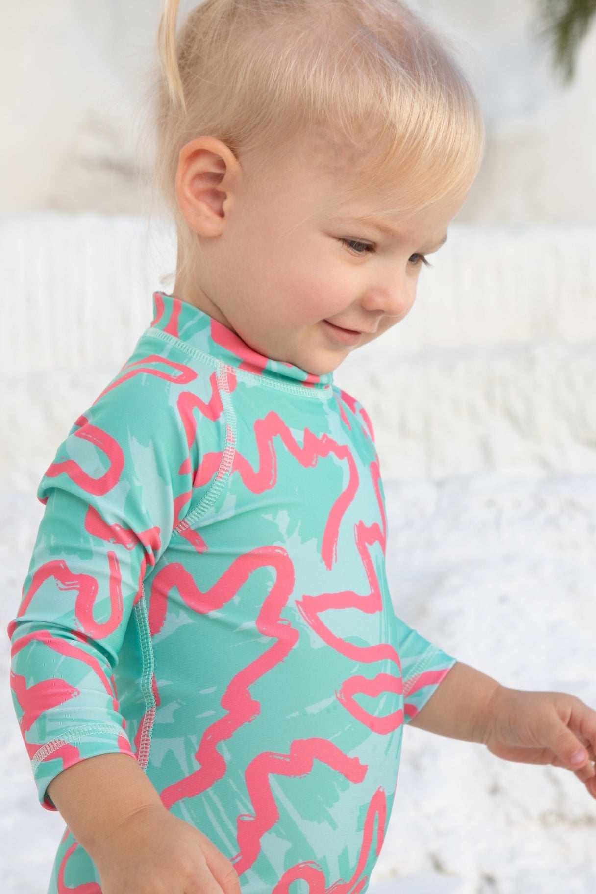 A young child with blonde hair wears the Baby Badeanzug - CORAL from kindischworld.com, standing outside against a white textured background with palm trees. This stylish swimsuit provides reliable UPF50+ sun protection.