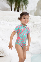 A young child in the Girls Badeanzug kurzarm - CORAL from kindischworld.com stands by a white stone pool with palm trees, smiling brightly in her colorful, patterned swimwear made from recycled polyester.
