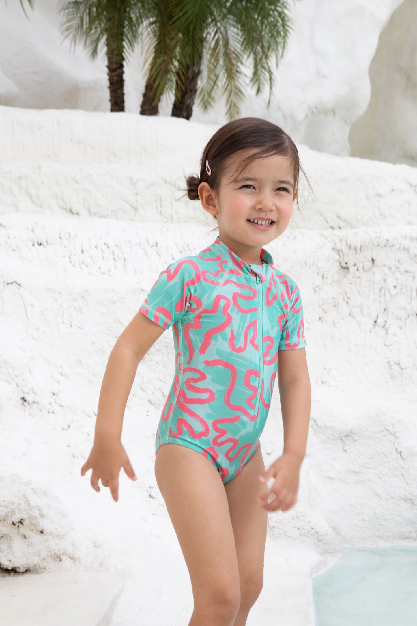 A young child in the Girls Badeanzug kurzarm - CORAL from kindischworld.com stands by a white stone pool with palm trees, smiling brightly in her colorful, patterned swimwear made from recycled polyester.