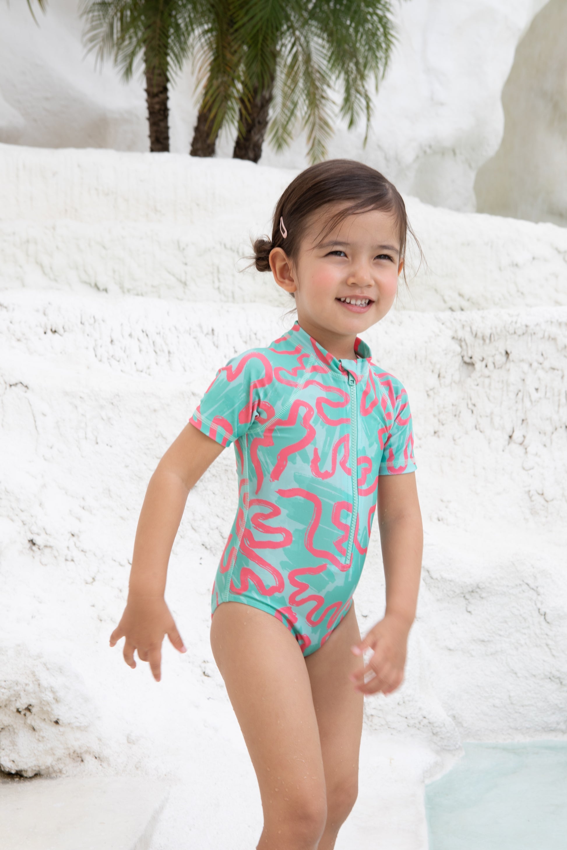 A young child in the Girls Badeanzug kurzarm - CORAL from kindischworld.com stands by a white stone pool with palm trees, smiling brightly in her colorful, patterned swimwear made from recycled polyester.