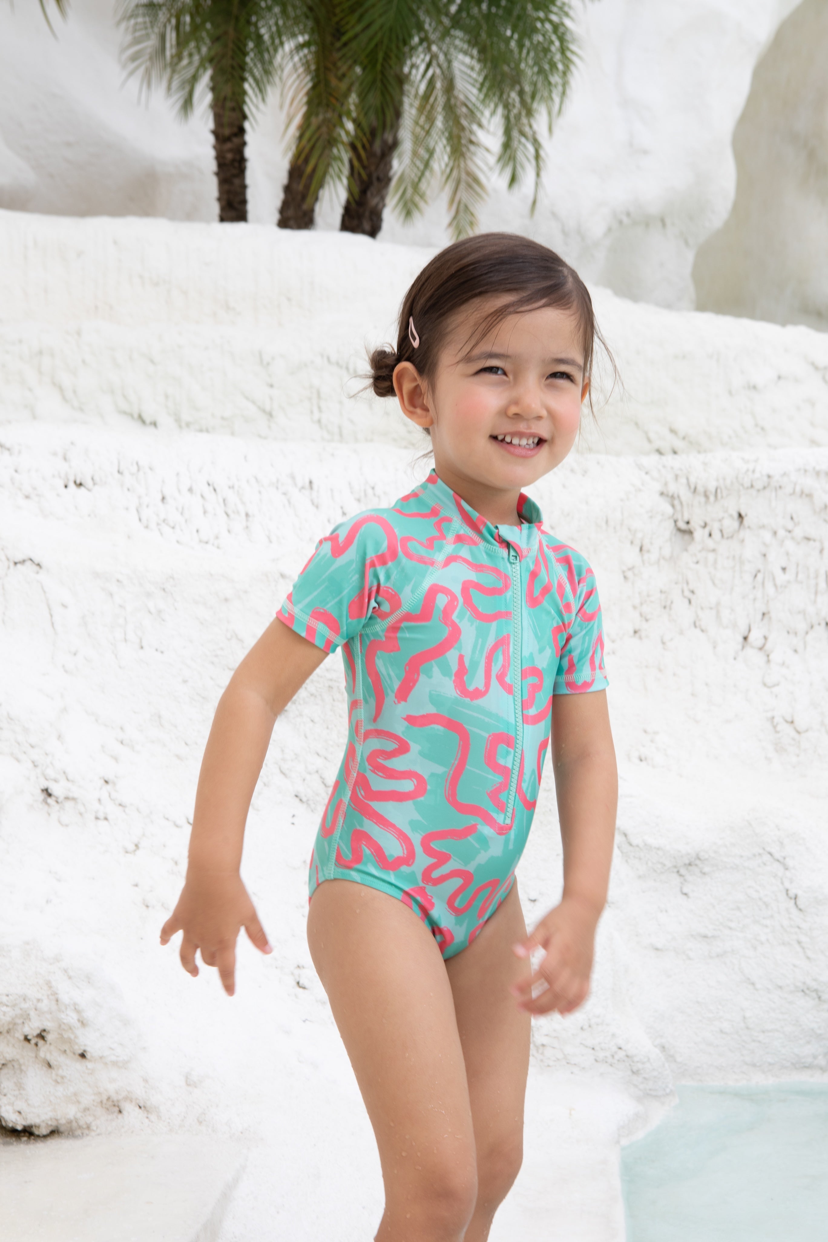 A young child in the Girls Badeanzug kurzarm - CORAL from kindischworld.com stands by a white stone pool with palm trees, smiling brightly in her colorful, patterned swimwear made from recycled polyester.