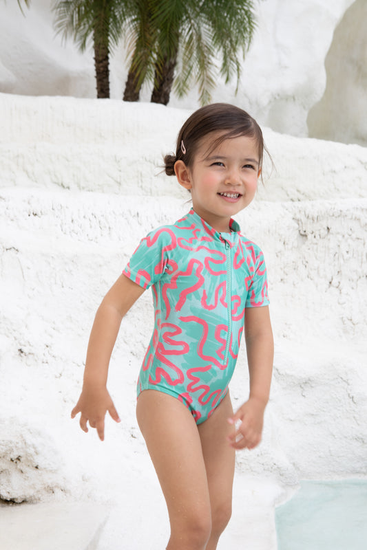 A young child in the Girls Badeanzug kurzarm - CORAL from kindischworld.com stands by a white stone pool with palm trees, smiling brightly in her colorful, patterned swimwear made from recycled polyester.