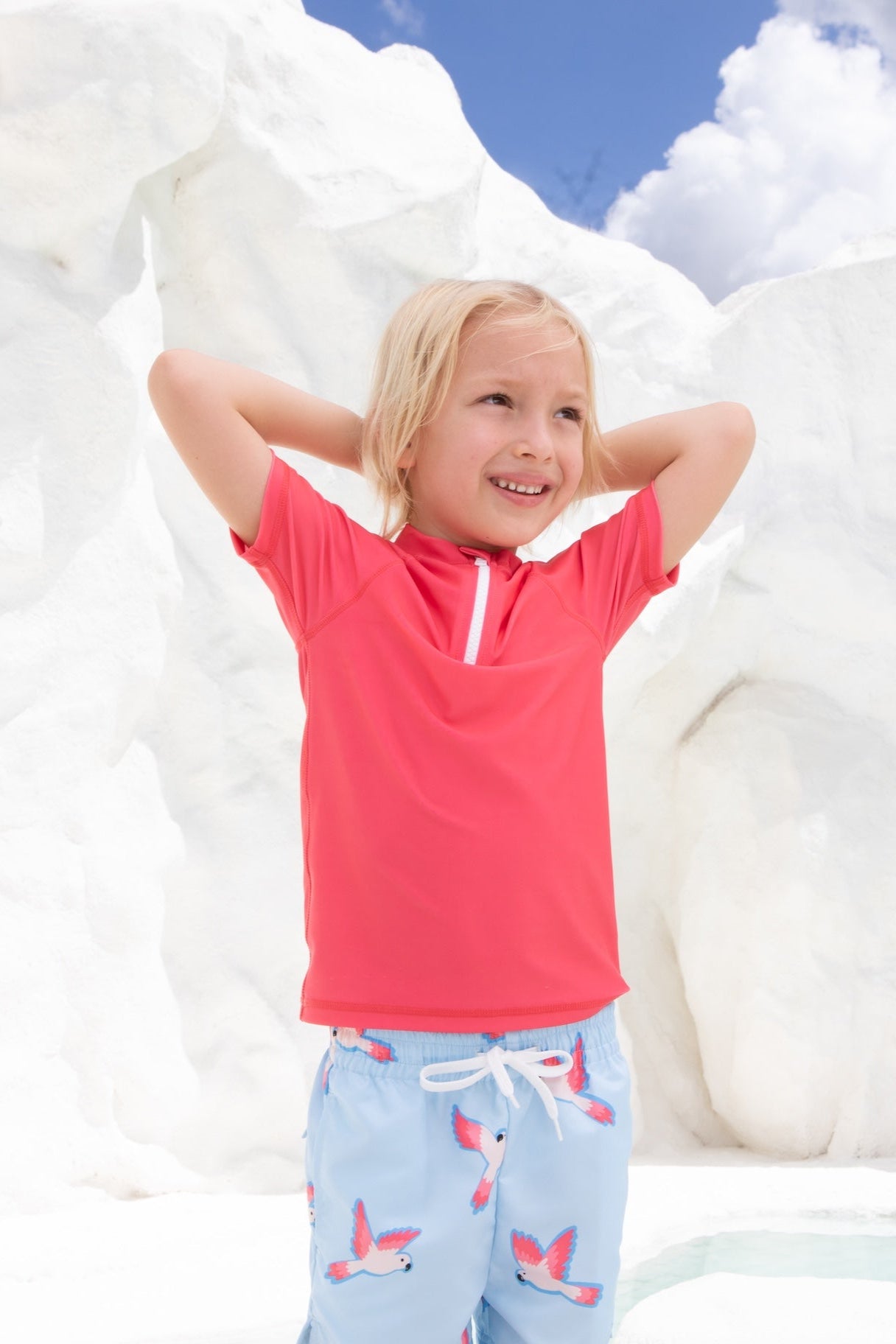 A smiling blond child stands with hands behind head, wearing the UV-Shirt kurzarm - Paradise Red by kindischworld.com and light blue swimwear with a bird print, set against bright white rocks under a partly cloudy sky.
