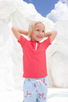 A smiling blond child stands with hands behind head, wearing the UV-Shirt kurzarm - Paradise Red by kindischworld.com and light blue swimwear with a bird print, set against bright white rocks under a partly cloudy sky.