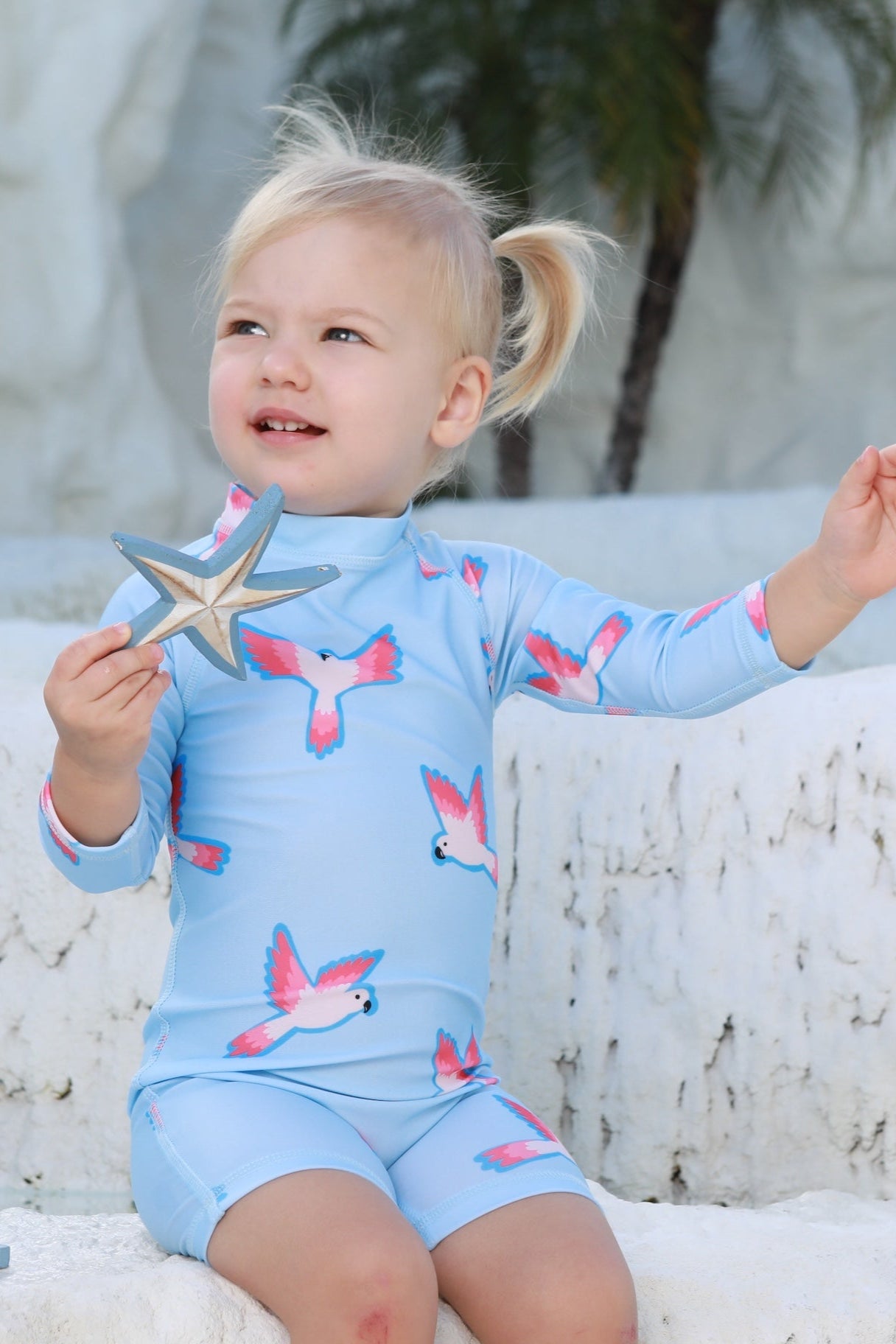 A young child with blonde pigtails sits on white stone steps, wearing the kindischworld.com Baby Badeanzug - PARROT (UPF50+) with bird prints, holding a star-shaped toy. A palm tree and beach ball are in the background.