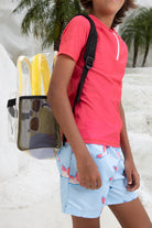 A child in a pink rash guard and blue parrot swim shorts stands by a white wall and pool, carrying the KINDISCH Kids Backpack - Yellow from kindischworld.com. Palm trees can be seen in the background.